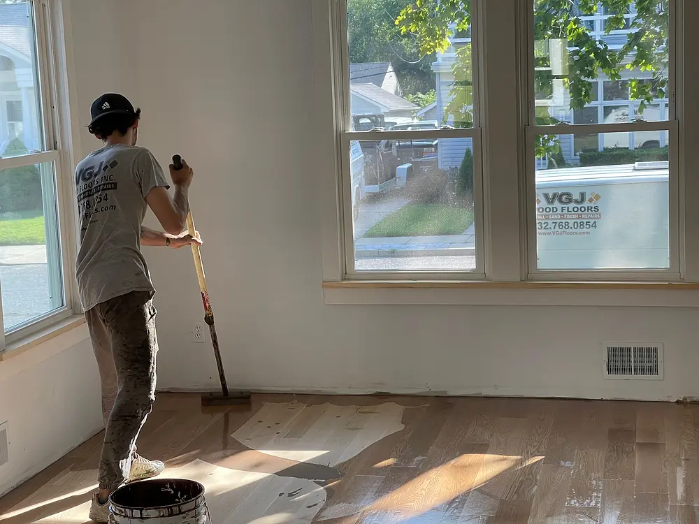 VGJ Floors crew member in a VGJ shirt applying finish, company van parked outside the window
