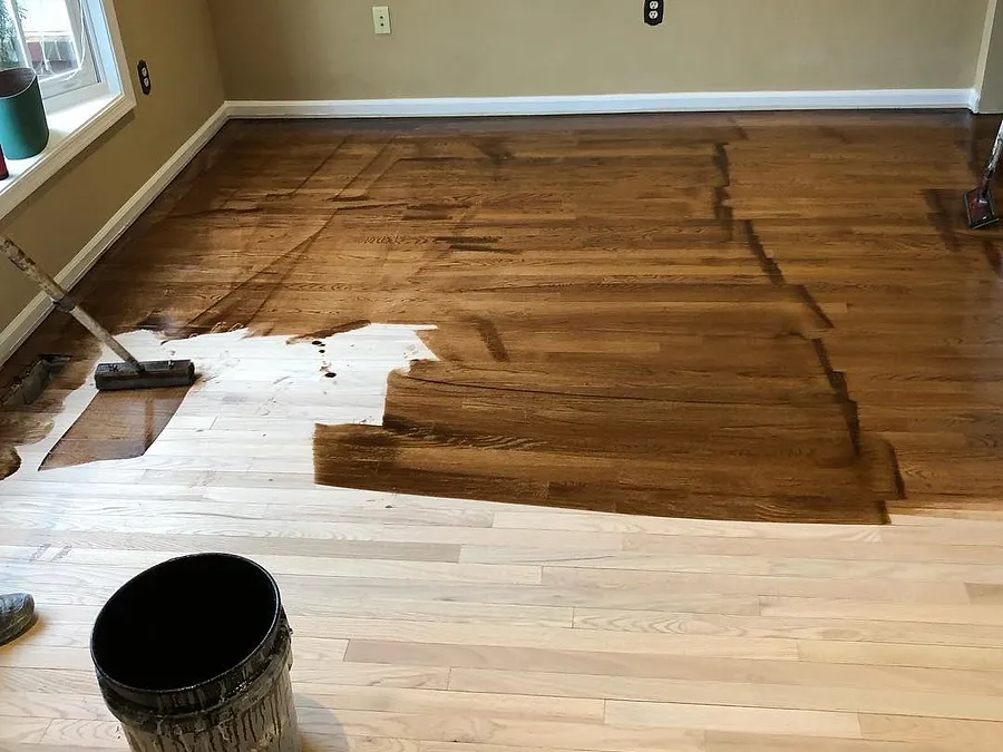 Dark stain being applied over freshly sanded hardwood, half the room done